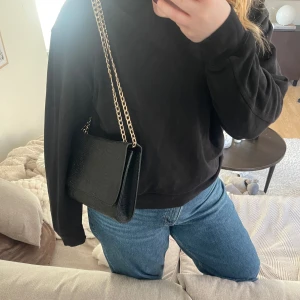 Svart axelväska med kedja - Snygg svart axelväska med guldfärgad kedja som axelrem. Väskan har en stilren design 