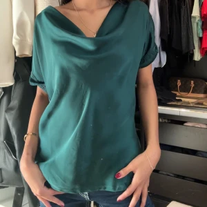 Grön blus med draperad hals - Snygg grön blus med draperad hals och korta ärmar. Perfekt för en stilren look. Blusen har en lös passform och är tillverkad i ett mjukt material som ger ett fint fall.