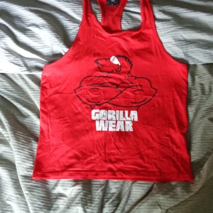 Röd linnetopp från Gorilla Wear - Säljer en röd linnetopp från Gorilla Wear med ett stort tryck av en muskulös gorilla på framsidan. Perfekt för träning och sommaraktiviteter. Linnet är i storlek M och har en sportig design.