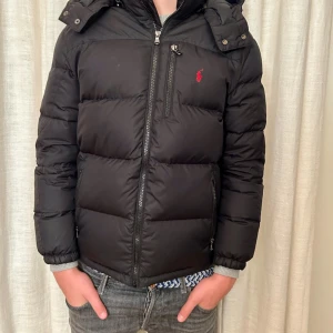 Svart dunjacka från Polo Ralph Lauren - Säljer en svart dunjacka från Polo Ralph Lauren med huva och dragkedja. Jackan har en broderad logotyp på bröstet och långa ärmar. Perfekt för kyliga dagar.