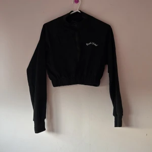Svart croppad jacka med dragkedja - Snygg svart croppad jacka med dragkedja framtill och textdetalj på bröstet. Perfekt för en avslappnad stil. Långärmad design med elastisk midja för en bekväm passform.