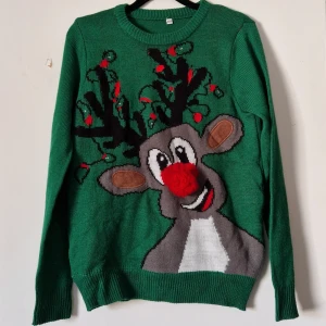 Grön jultröja med renmotiv - Säljer en grön stickad jultröja från Maximus med ett stort renmotiv på framsidan. Tröjan har långa ärmar och är perfekt för julfirandet. Den är dekorerad med röda och svarta detaljer som ger en festlig känsla.