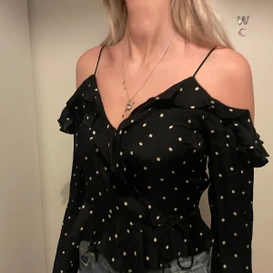 Svart prickig offshoulder topp - Snygg svart offshoulder topp med vita prickar och volangdetaljer. Perfekt för en trendig look med smala axelband och långa ärmar.