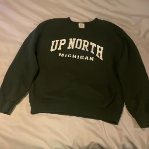 Mörkgrön sweatshirt med tryck - Säljer en mörkgrön sweatshirt med texten 'UP NORTH MICHIGAN' i vitt på framsidan. Tröjan har långa ärmar och en klassisk rund halsringning. Perfekt för en avslappnad stil.