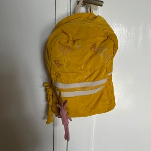 Gul ryggsäck med broderad text - Snygg gul ryggsäck med broderad text på framsidan. Väskan har en dragkedja upptill och en praktisk ficka framtill. Perfekt för att bära med sig det nödvändigaste. En rosa nyckelring hänger på sidan för extra stil.