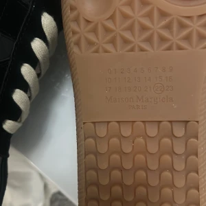  Maison Margiela gats - Snygga svarta sneakers från Maison Margiela med beige sula och vita snören. Skorna har en stilren design med en kombination av skinn och mocka. I 44