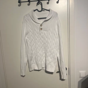 Vit stickad tröja med knappar - Säljer en stilren vit stickad tröja med ribbad krage och två knappar framtill. Tröjan har ett snyggt flätmönster och långa ärmar, perfekt för kyligare dagar.