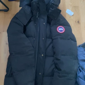 Svart dunjacka från Canada Goose - Säljer en svart dunjacka från Canada Goose med huva och dragkedja. Jackan har en broderad logotyp på bröstet och är perfekt för kalla vinterdagar. Den är långärmad och har en klassisk design. Varan är äkta och tar även emot byten:)