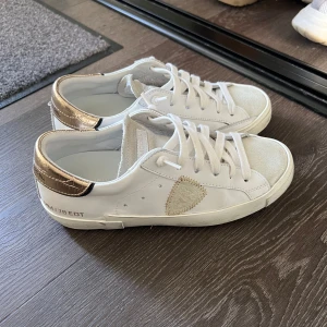 Vita sneakers från Philippe Model - Snygga vita sneakers från Philippe Model med gulddetaljer på hälen och sidan. Skorna har snörning och en unik design med en textur på sidan. Perfekta för en stilren look. Skokartong finns kvar men inte dustbag. Har en liten defekt som visas på sista bilden och vid hälen, men inget som man ser. 
