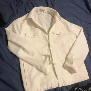 Vit jacka från Zara - Snygg vit jacka från Zara med klassisk krage och knappar framtill. Jackan har två bröstfickor med lock och knappar, vilket ger en stilren look. Perfekt för vår och sommar. Köpte den för ungefär 3 månader sen för ca 850kr, säljer den pga att de inte är min stil längre.den ser ut så för den har varit i tvätten en gång, men det går att få den se ny ut med strykjärn 