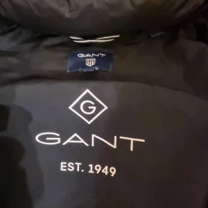 Svart dunjacka från GANT - Snygg svart dunjacka från GANT med dragkedja och avtagbar huva. Jackan har en quiltad design och är perfekt för kalla dagar. Märkeslogga på bröstet ger en stilren touch.