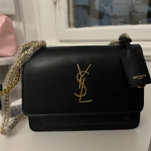 Svart axelväska från Saint Laurent - Elegant svart axelväska från Saint Laurent i skinn med guldfärgad YSL-logga på framsidan. Väskan har en kedjerem och ett magnetiskt knäppe. Perfekt för att ge en lyxig touch till din outfit.