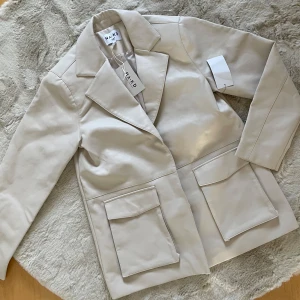 Beige kavaj från NA-KD 36 oversize oanvänd  - Snygg beige kavaj från NA-KD med klassisk design. Den har långa ärmar och stora fickor framtill. Perfekt för en stilren look.