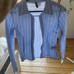 Blå och vit randig figursydd skjorta - Snygg blå och vit randig skjorta med knappar framtill. Perfekt för en stilren look. Skjortan har långa ärmar och en klassisk krage. Sitter jättesnygg åt sittande 