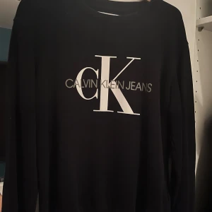 Svart långärmad tröja från Calvin Klein - Snygg svart långärmad tröja från Calvin Klein med stort CK-logotyptryck på framsidan. Perfekt för en stilren och avslappnad look.