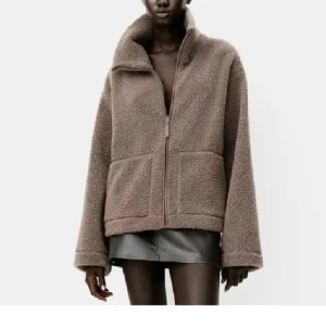 Beige teddyjacka med dragkedja - Mysig beige teddyjacka med dragkedja framtill och stora fickor. Perfekt för kyliga dagar med sin fluffiga och varma design. Jackan har en hög krage och långa ärmar för extra komfort.