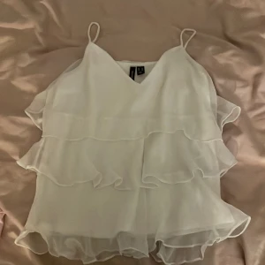 Vit volangtopp från Vero Moda - Säljer en söt vit volangtopp från Vero Moda. Toppen har tunna axelband och flera lager av volanger som ger en luftig och romantisk känsla. Perfekt för varma sommardagar eller en festlig kväll!💗