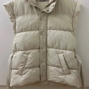 Beige dunväst med bälte - Snygg beige dunväst med bälte i midjan. Perfekt för kyligare dagar. Den är endast testad. Säljer eftersom att den är för stor för mig, storlek S men skulle säga att den är stor i storleken och skulle sitta bättre på någon som har storlek M eller L❣️
