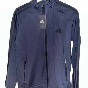 Svart träningskofta från Adidas - Snygg svart träningsjacka från Adidas med dragkedja och de klassiska tre ränderna längs ärmarna. Jackan har en hög krage och två praktiska fickor med dragkedja framtill. Perfekt för träning eller för dagligen 