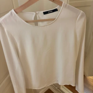Vit långärmad topp/blus från Bik Bok - Säljer en stilren vit långärmad topp/blus från Bik Bok. Toppen har en rund halsringning och en knappdetalj i nacken. Perfekt för en minimalistisk look. Skönt och fint material! Använd fåtal gånger!