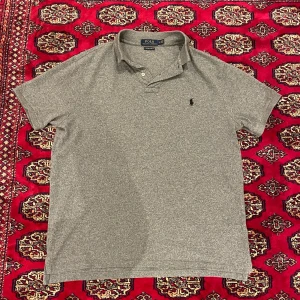 Grå pikétröja från Ralph Lauren - Säljer en klassisk grå pikétröja från Ralph Lauren med korta ärmar och en liten broderad logotyp på bröstet. Perfekt för en stilren och avslappnad look. Nästan helt ny använd ca 5-6 gånger. Storlek L men sitter mindre!                                                                          Vid frågor och funderingar hör gärna av er🤗                    Mvh Doré.reselll