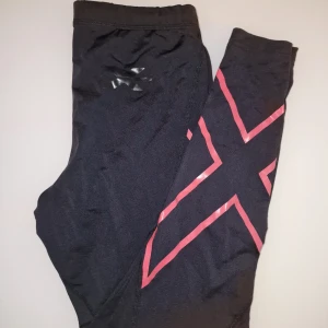 Svarta kompressionstights från 2xU - Snygga svarta träningsleggings med rosa mönster och logga från märket 2xU. Kompressionstights hjälper till att minska risken för skador och påskynda återhämtningen så att du kan fortsätta att prestera på topp. Liten defekt på 2 bilden då färgen lossnat längst ner på märket. 