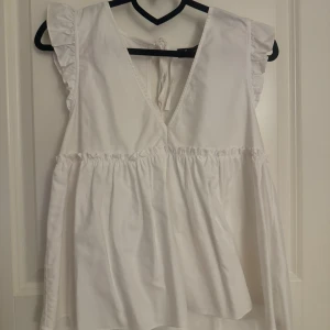 Vit blus med volangdetaljer - Zara - Säljer en söt vit blus med volangdetaljer på axlarna och en v-ringad design. Blusen har en knytning i ryggen och är perfekt för en somrig look.