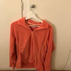 Orange fleecejacka från Line One - Mysig orange fleecejacka från Line One med dragkedja framtill. Perfekt för kyliga dagar. Jackan har en enkel design med långa ärmar och en hög krage.