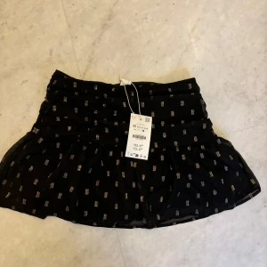 Svart kortkjol med guldmönster - Svart kortkjol med ett guldmönster. Kjolen har en volangdetalj och dragkedja i sidan för enkel påklädning. Perfekt för sommaren och lite finare tillställningar. Kjolen är helt ny och oanvänd. Nypris 450kr