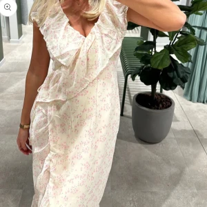 Blommig klänning med volangdetaljer - Klänning i ljus färg med små blommor och volangdetaljer. Klänningen har en omlott-design. Jag har endast använt klänningen en gång på ett bröllop så den är i väldigt fint skick. 