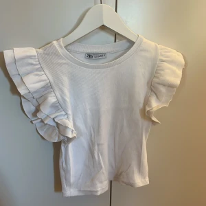 Vit ribbad topp med volangärmar från Zara - Säljer en söt vit ribbad topp från Zara med volangdetaljer på ärmarna. Perfekt för en somrig look. Toppen har en rund halsringning och är i ett mjukt material.