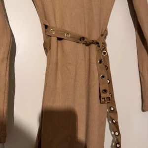 Beige klänning från Missguided - Snygg beige klänning från Missguided med ribbad textur och ett bälte med nitar. Klänningen har långa ärmar och en figurnära passform. Perfekt för en stilren look.