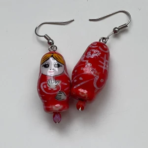 Röda örhängen med dockmotiv - Charmiga röda örhängen med handmålade dockmotiv i porslin De har en unik design med små detaljer i vitt och svart samt en liten pärla längst ner. Perfekta för att ge en färgglad touch till din outfit.