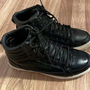 Svarta sneakers från Marine Classics fodrade stl 43 - Snygga svarta sneakers med foder från Marine Classics med hög snörning och en vit sula. Skorna har en stilren design med ett emblem på sidan. Perfekta för en casual look.