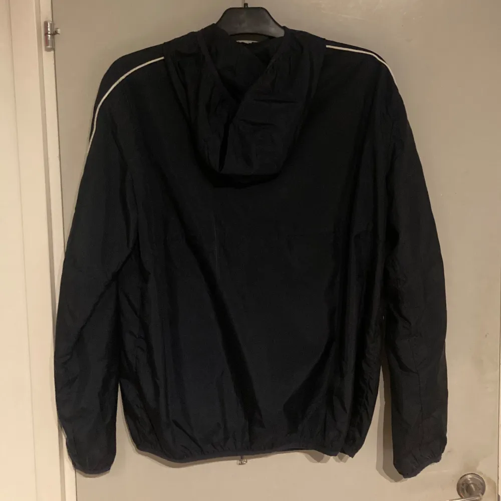 Snygg mörkblå vindjacka från Armani Exchange med dragkedja och huva. Jackan har vita detaljer längs ärmarna och logotypen A|X på bröstet. Perfekt för en vår och sommar. Priset är diskuterbart. Takit.