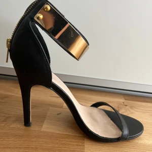 Svarta klackskor med gulddetaljer - Eleganta svarta klackskor med en smal rem över tårna och en bred guldfärgad rem runt ankeln. Skorna har en dragkedja baktill och en spetsig tå. Perfekta för en stilfull look.