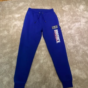 Blå mjukisbyxor från Hoodrich - Snygga blå mjukisbyxor från Hoodrich med vit och röd logga på benet. Byxorna har snörning i midjan och ribbade muddar vid bensluten för en bekväm passform. Perfekta för en avslappnad stil.