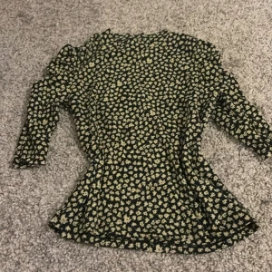 Blommig topp med volangdetaljer - Snygg blommig topp med volangdetaljer på axlarna. Toppen har en figurnära passform och trekvartsärmar. Perfekt för att ge en romantisk touch till din outfit.