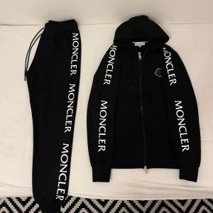 Moncler tracksuit svart - Snygg svart hoodie och matchande byxor från Moncler med vit logotyp längs ärmarna och benen. Hoodien har dragkedja och justerbar huva. Perfekt för en stilren och bekväm look. Helt nya använt 3 gånger