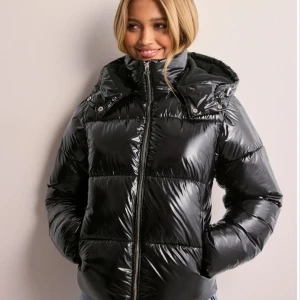 Svart glansig pufferjacka - Snygg svart pufferjacka med glansig finish och dragkedja framtill. Jackan har en hög krage och huva för extra värme. Perfekt för kyliga dagar. Säljer den då jag har köpt en annan men använde den inte alls många gånger, i super skick dessutom!