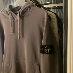 Grå hoodie från Stone Island - Säljer en stilren grå hoodie från Stone Island med klassisk logga på ärmen. Tröjan har en bekväm passform med justerbar huva och känguruficka framtill. Perfekt för en avslappnad look. Tröjan är i storlek S köpt från NK i Göteborg 