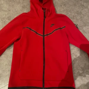  nike tech hoodie - En snygg röd nike tech! Ett pyttelitet slitning/hål vid dragkedjan på magen, skriv för bild! Säljs då jag ej använder!
