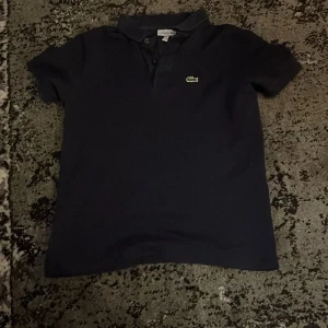 Mörkblå pikétröja från Lacoste - Säljer en klassisk mörkblå pikétröja från Lacoste med den ikoniska krokodilloggan på bröstet. Tröjan har korta ärmar och en krage med knappar. Perfekt för en stilren och avslappnad look.