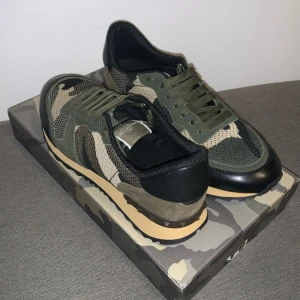 Valentino Garavani sneakers i camouflage - Snygga Valentino Garavani sneakers med ett coolt camouflage-mönster. Skorna har snörning och en kombination av gröna och svarta detaljer. Perfekta för en trendig look. 2 slitningar på lådan men inget stort, Passar även bra på storlek 44