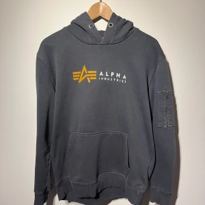 Grå hoodie från Alpha Industries - Säljer en snygg grå hoodie från Alpha Industries med deras ikoniska logga i gult på bröstet. Tröjan har en praktisk ficka framtill och en ärmficka med dragkedja. Perfekt för en casual look. Inköpt på Carlings 