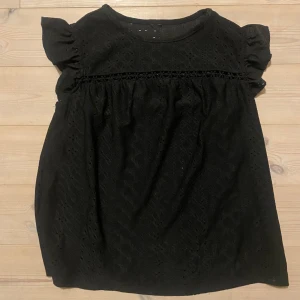 Svart blus med spetsdetaljer - Snygg svart blus i storlek XS med spetsdetaljer och korta volangärmar. Perfekt för en stilren look. Blusen har en rund hals och ett diskret mönster som ger en elegant touch.
