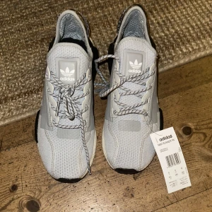 Grå Adidas NMD R1 sneakers - Snygga grå Adidas NMD R1 sneakers med vit sula och svarta detaljer. Skorna har en stickad ovandel och klassisk snörning. Perfekta för en stilren och bekväm look.