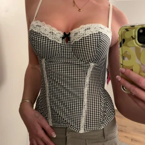 Svartvit rutig korsett med spetsdetaljer - Snygg korsett i svartvitt rutigt mönster med spetsdetaljer och en liten rosett framtill. Den har smala axelband och en figurnära passform som framhäver midjan. Perfekt för en trendig look!
