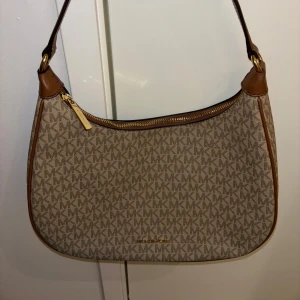 Beige/brun axelväska från Michael Kors - Snygg beige axelväska från Michael Kors med klassiskt MK-mönster. Väskan har bruna läderdetaljer och en praktisk dragkedja upptill. Inga skavanker och aldrig använd. Denna väska är perfekt för både vardagsbruk och festliga tillfällen, och lägger till en touch av elegans till vilken outfit som helst. Passa på att förnya din stil med denna tidlösa accessoar! Kontakta mig för mer information eller bilder.