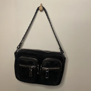 Svart axelväska från Noella - Snygg svart axelväska från Noella med två framfickor och dragkedjor. Väskan har en justerbar axelrem med nitar för extra stil. Perfekt för att bära dina nödvändigheter med stil. Jätte lite luddig men va så när jag fick den.
